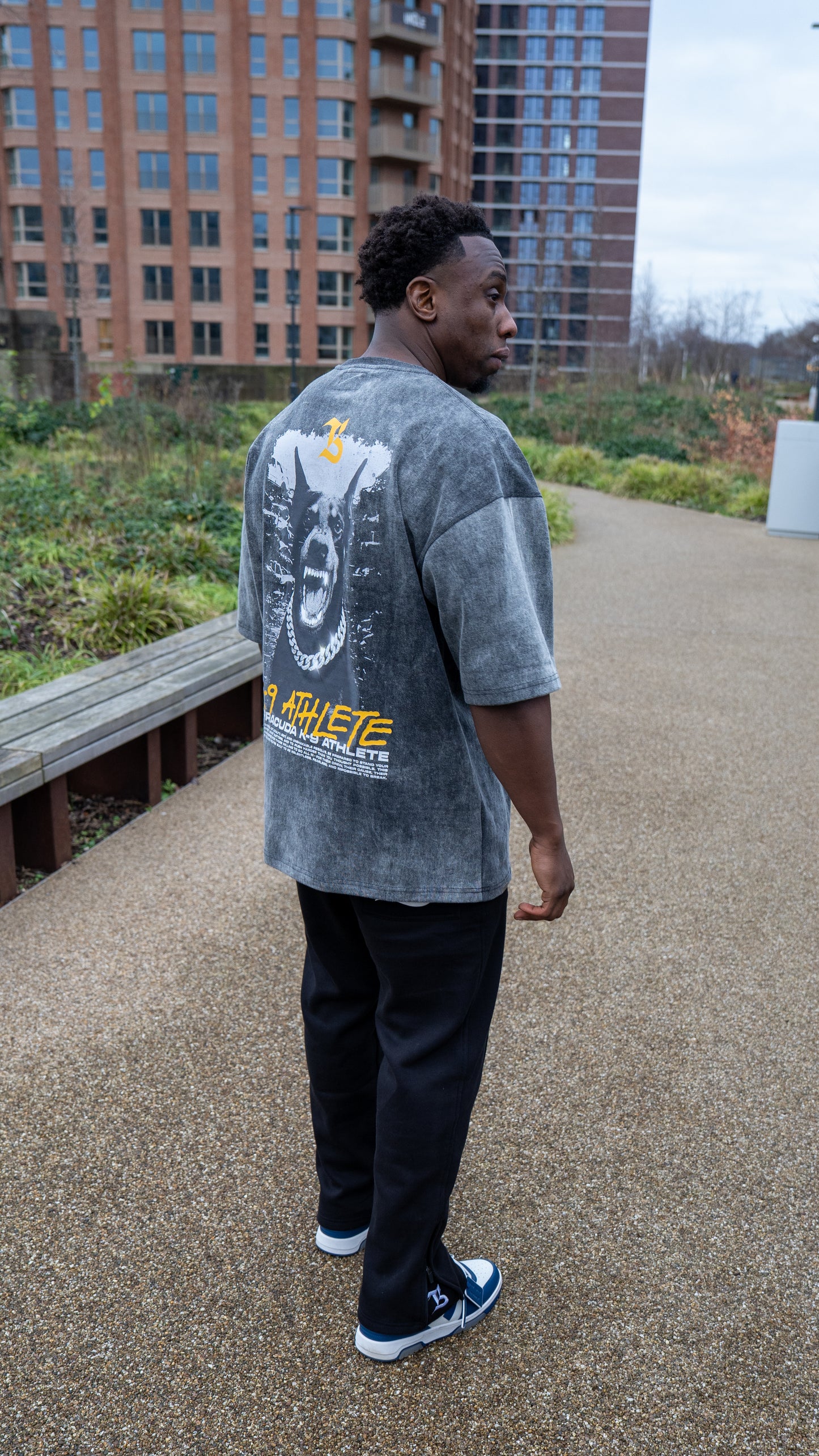 Person wearing a gray t-shirt with a graphic design, standing on a pathway with buildings in the background.