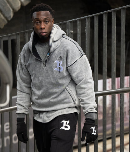 Person wearing a gray hoodie and black pants with logos, standing against a metal railing.