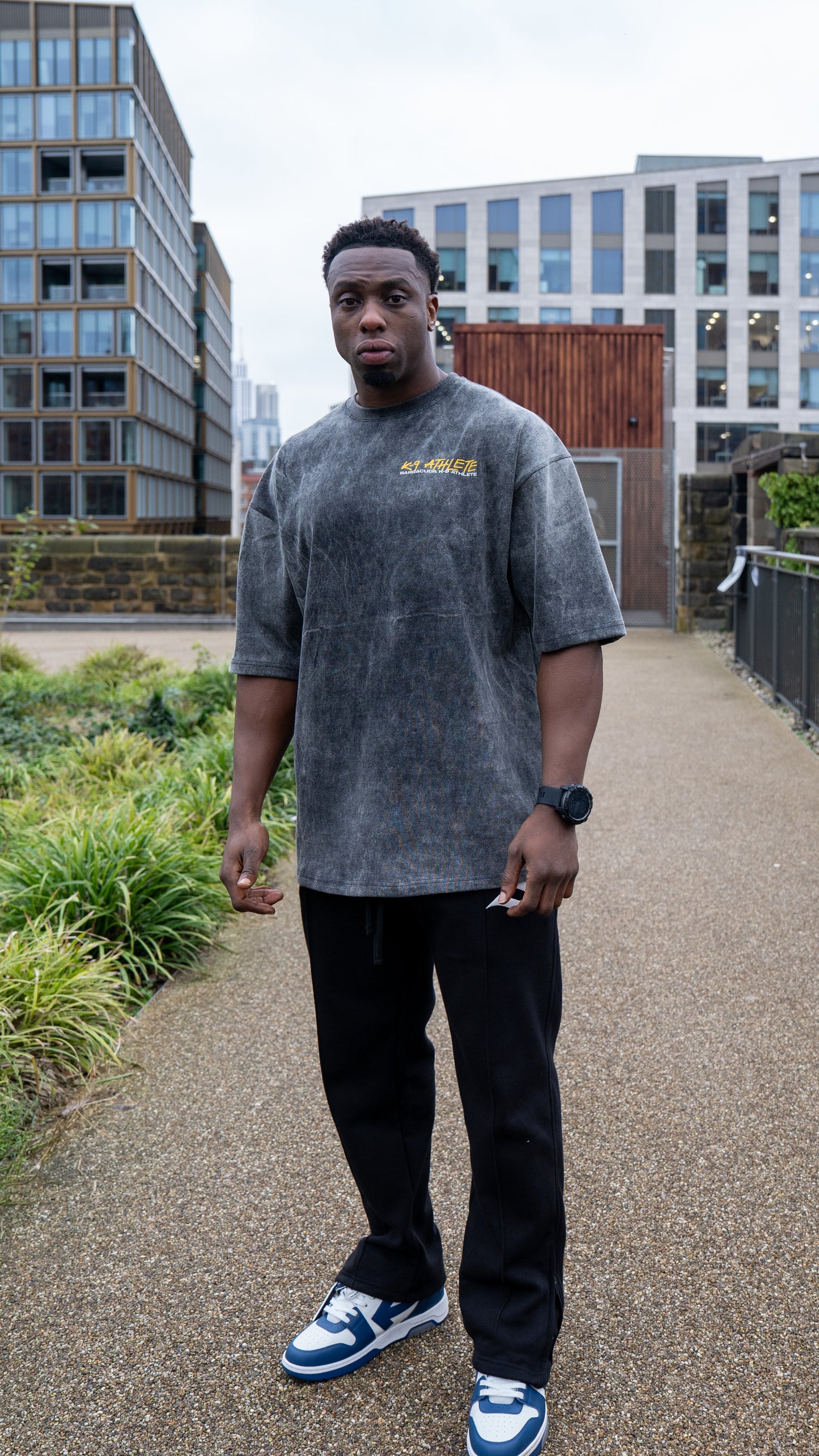 Person wearing a grey t-shirt with text and black pants standing on a path with buildings in the background