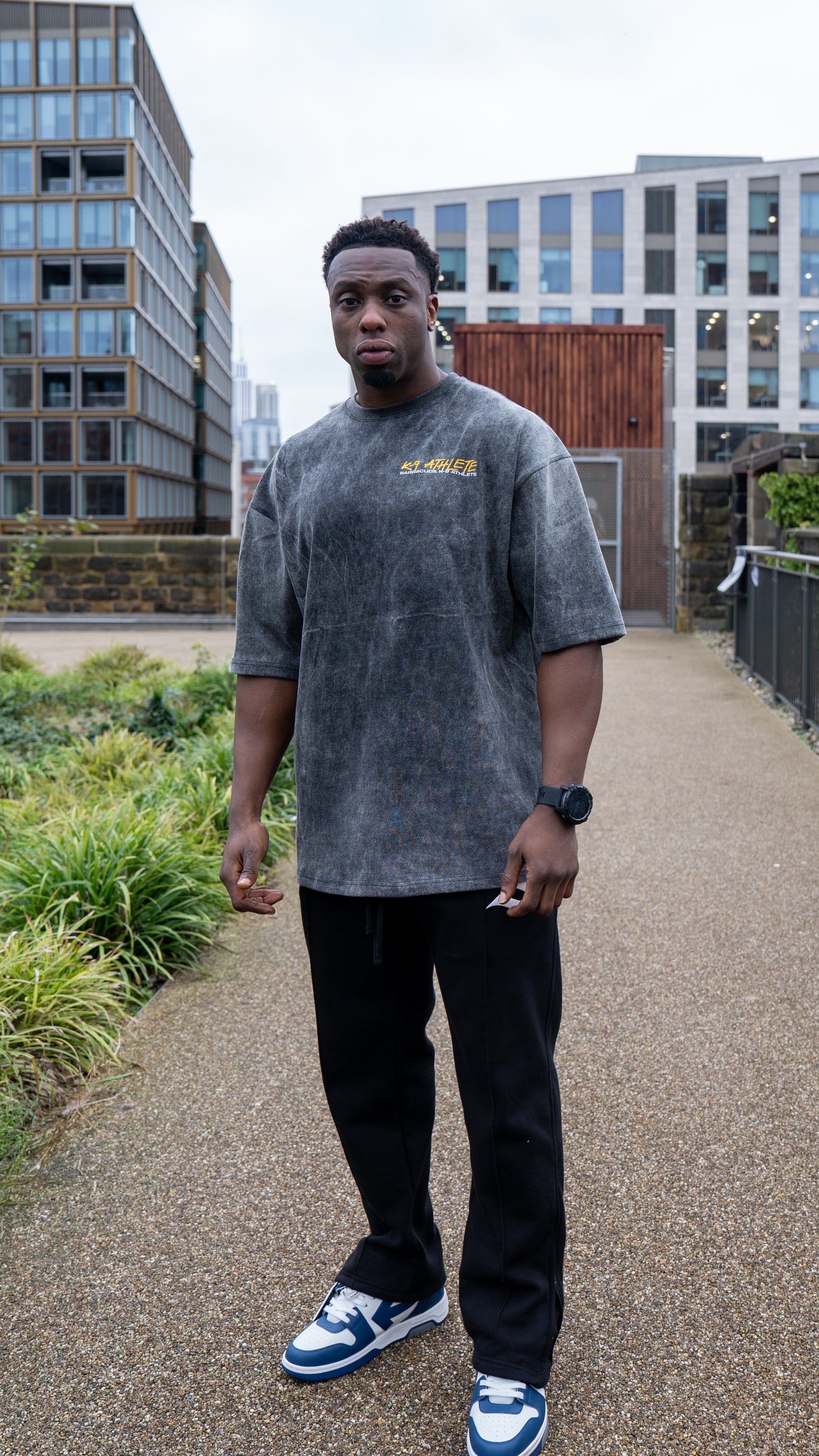 Person wearing a grey t-shirt with text and black pants standing on a path with buildings in the background