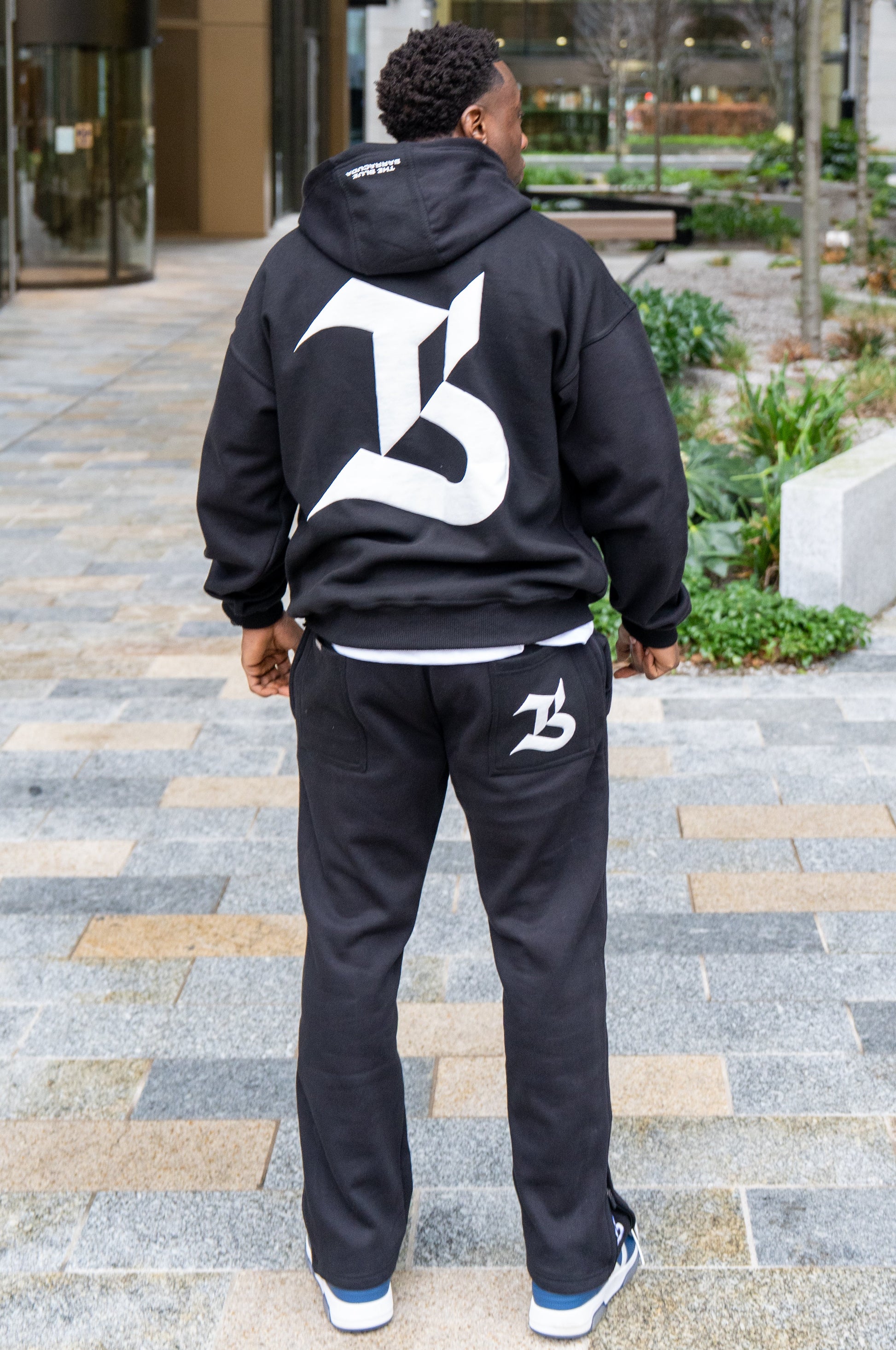 Person wearing a black hoodie and pants with a logo, standing on a paved area.