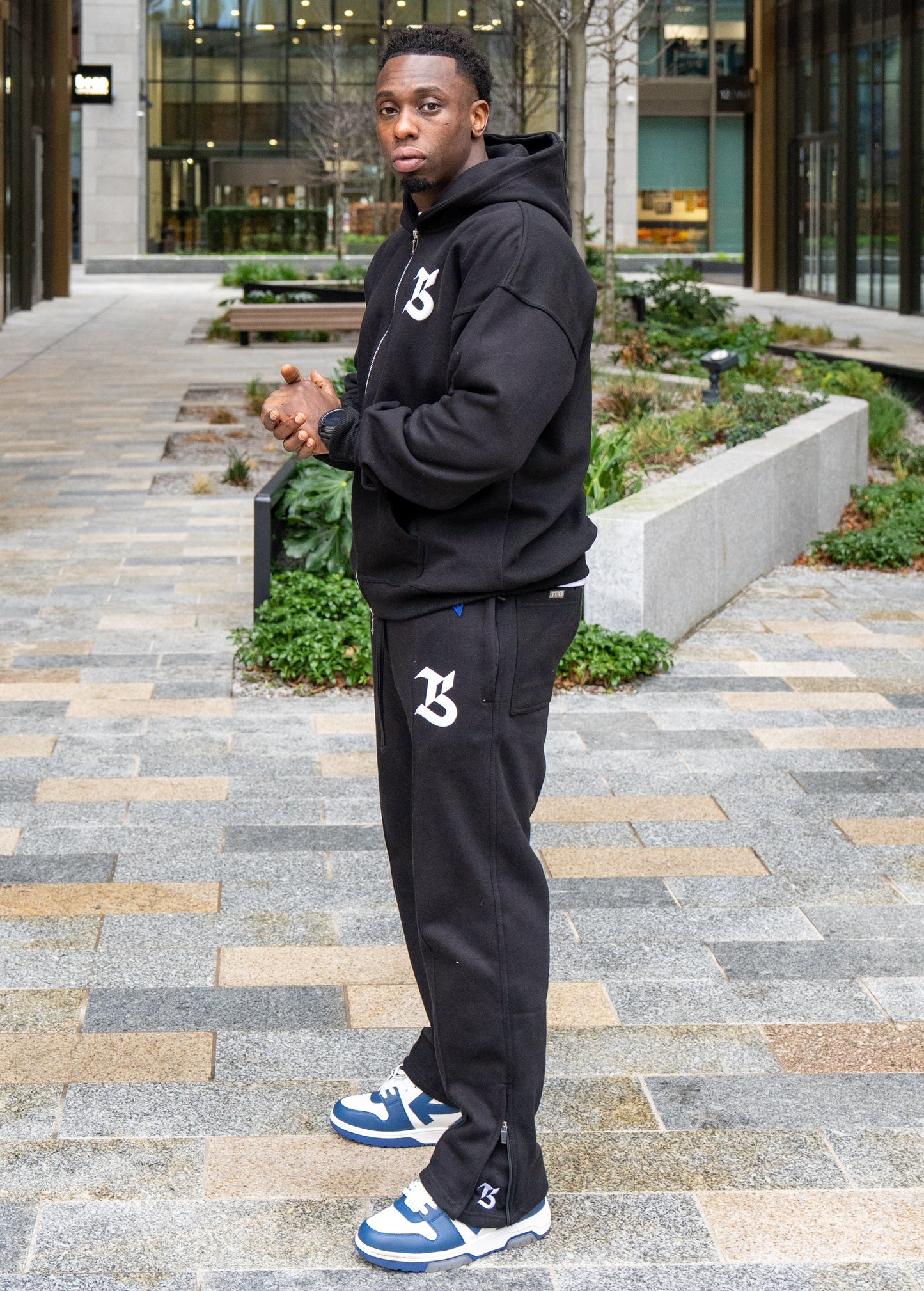 Person wearing a black hoodie and pants with a logo, standing on a paved walkway.
