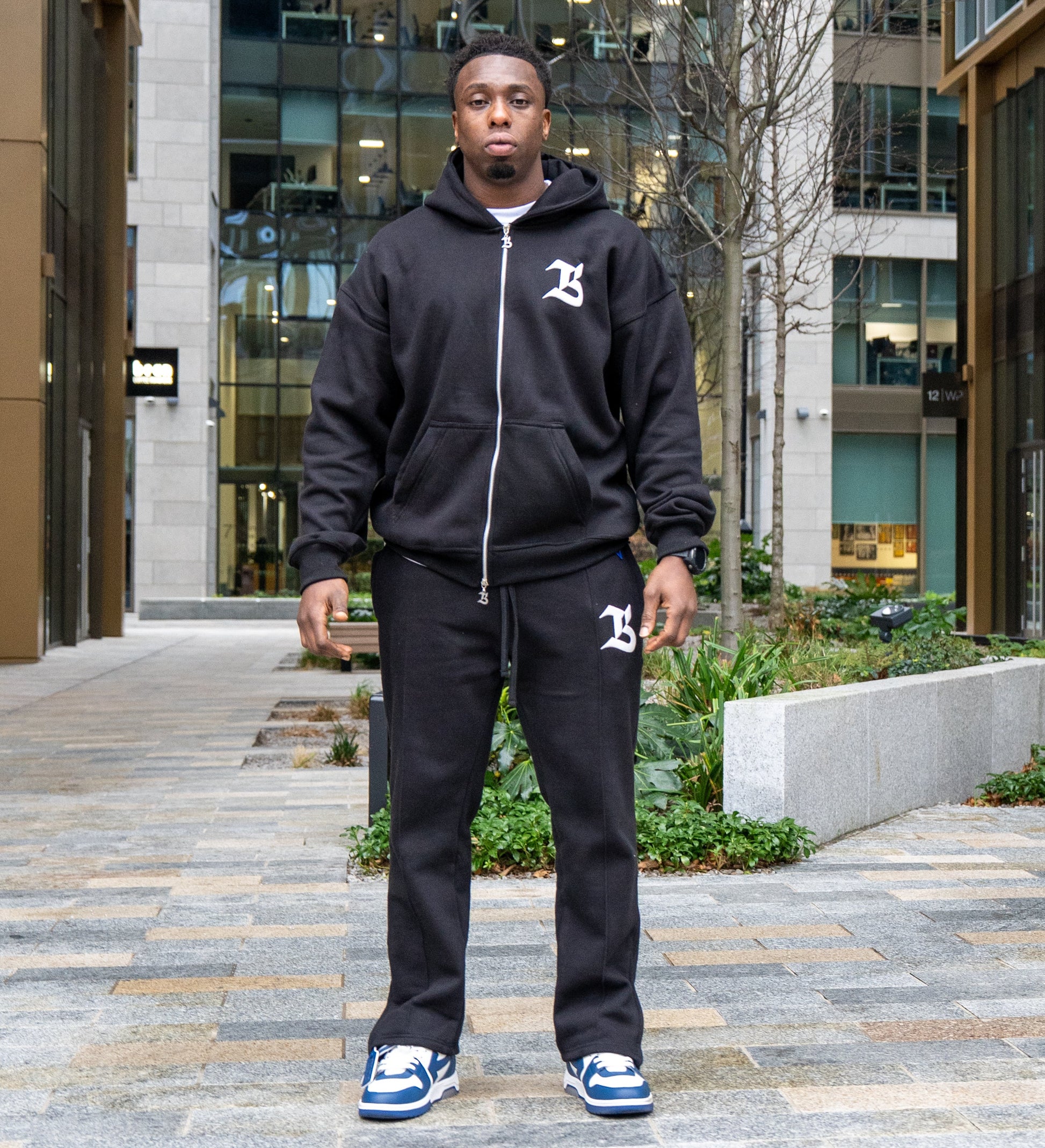 Person wearing a black hoodie and pants with a logo, standing on a city street.