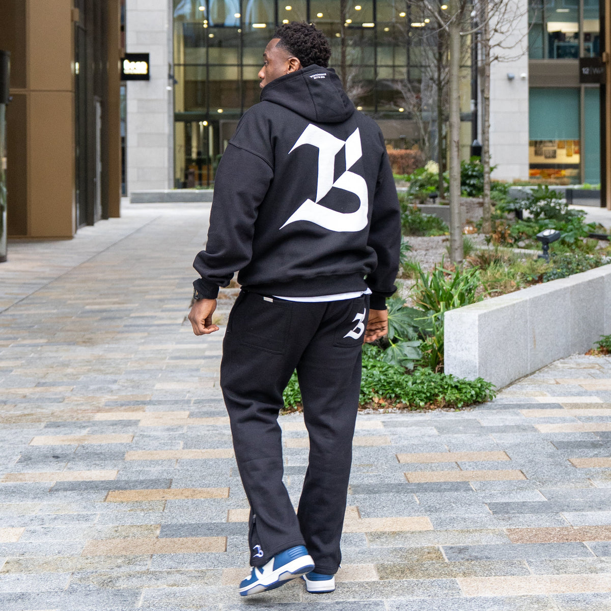 Person wearing a black hoodie with a large white letter 'B' walking on a sidewalk.