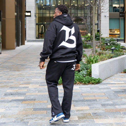 Person wearing a black hoodie with a large white letter 'B' walking on a sidewalk.